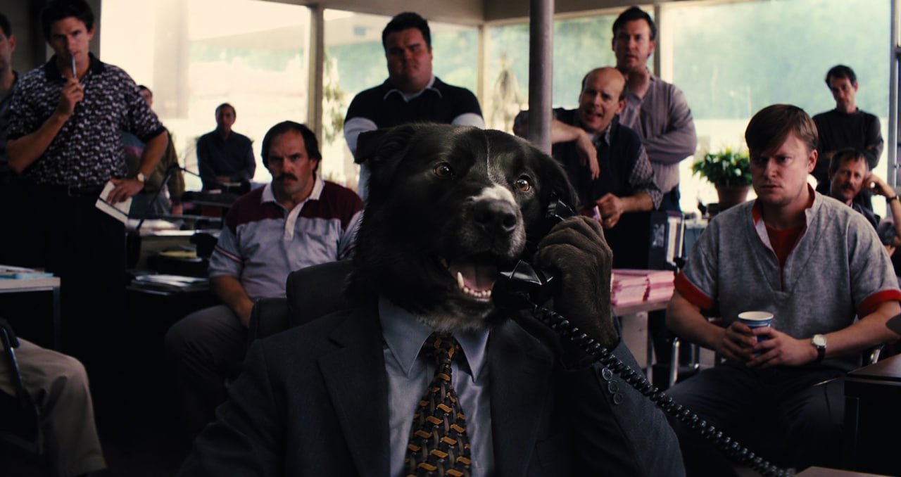 A black Labrador in a suit and tie sits behind a desk holding a telephone to his ear while a room full of office workers looks on.